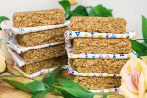 Two stacks of flapjacks with a white and triangle coloured cloth separating each bar on a wooden board surrounded with flowers
